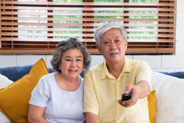 Older couple sitting on couch watching TV. Man is pointing the remote at the TV.