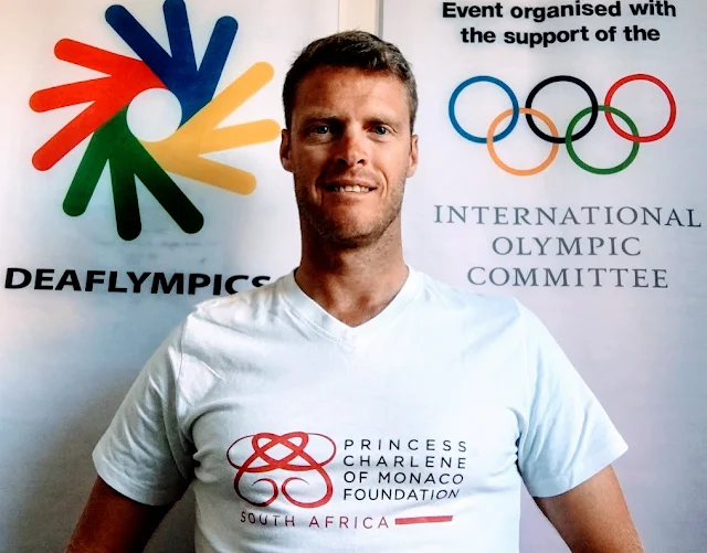 athlete standing in front of Deaflympics and International Olympic Committee logos wearing foundation t-shirt