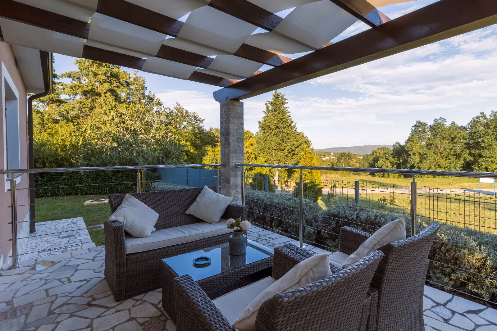 Outdoor patio with wicker furniture under a pergola, overlooking a green landscape with trees and grass.