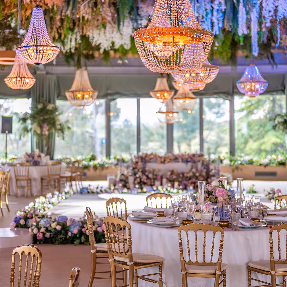 Sofisticado salón de bodas con mesas redondas decoradas con arreglos florales y candelabros de cristal colgantes.