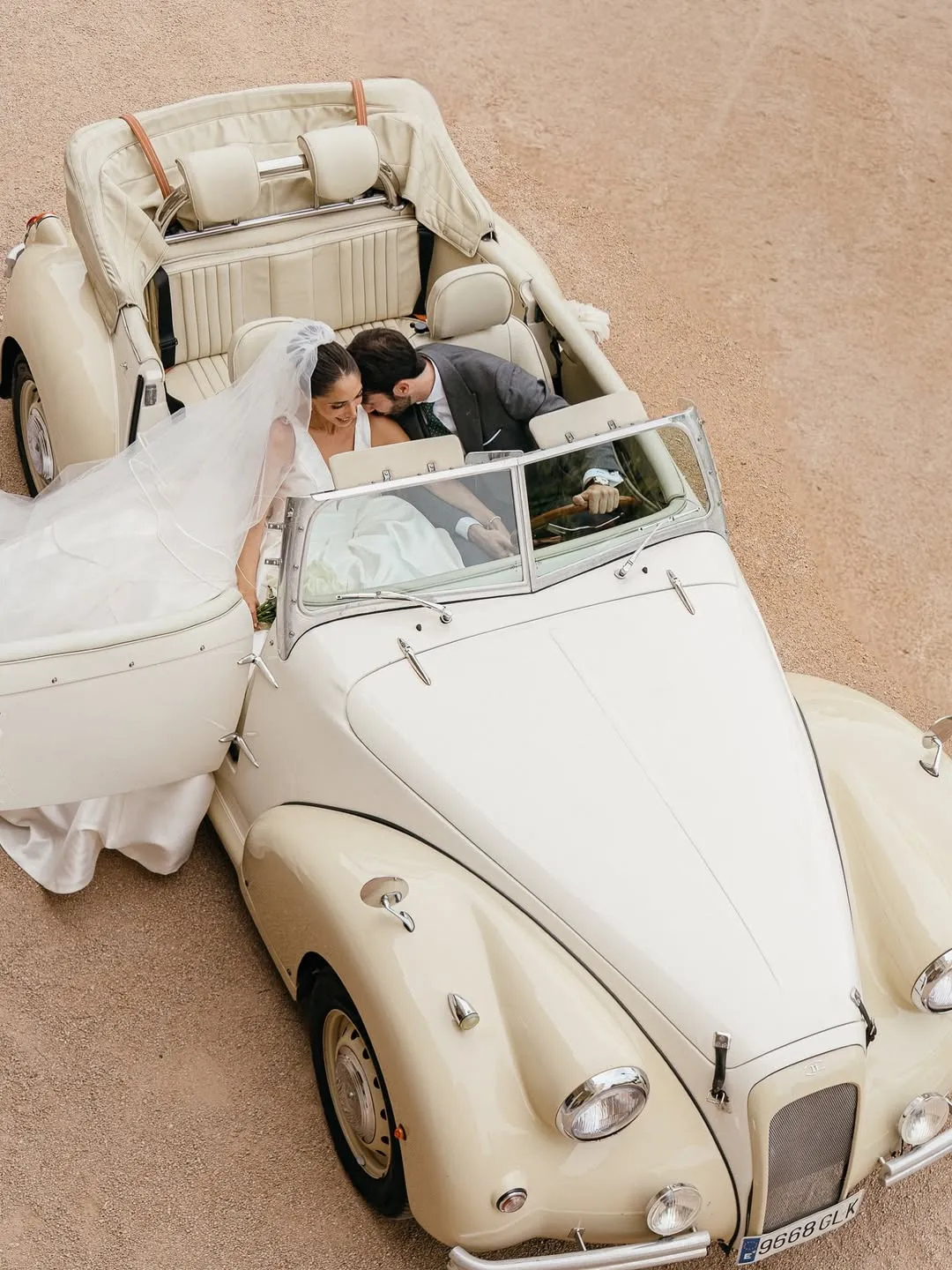 Pareja de recién casados sonrientes sentados en un coche antiguo blanco descapotable con la puerta abierta.
