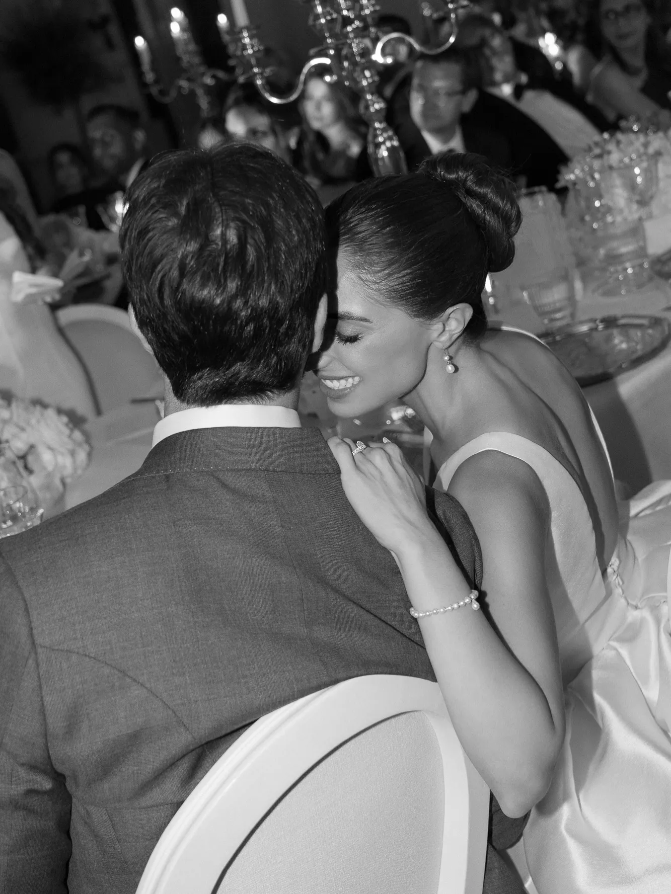 Pareja abrazada sonriente en una boda, ella con vestido y joyas, él de espaldas con traje.