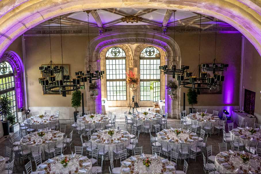 Salón de armas con mesas redondas decoradas para un evento, con sillas blancas, arreglos florales y luces moradas.