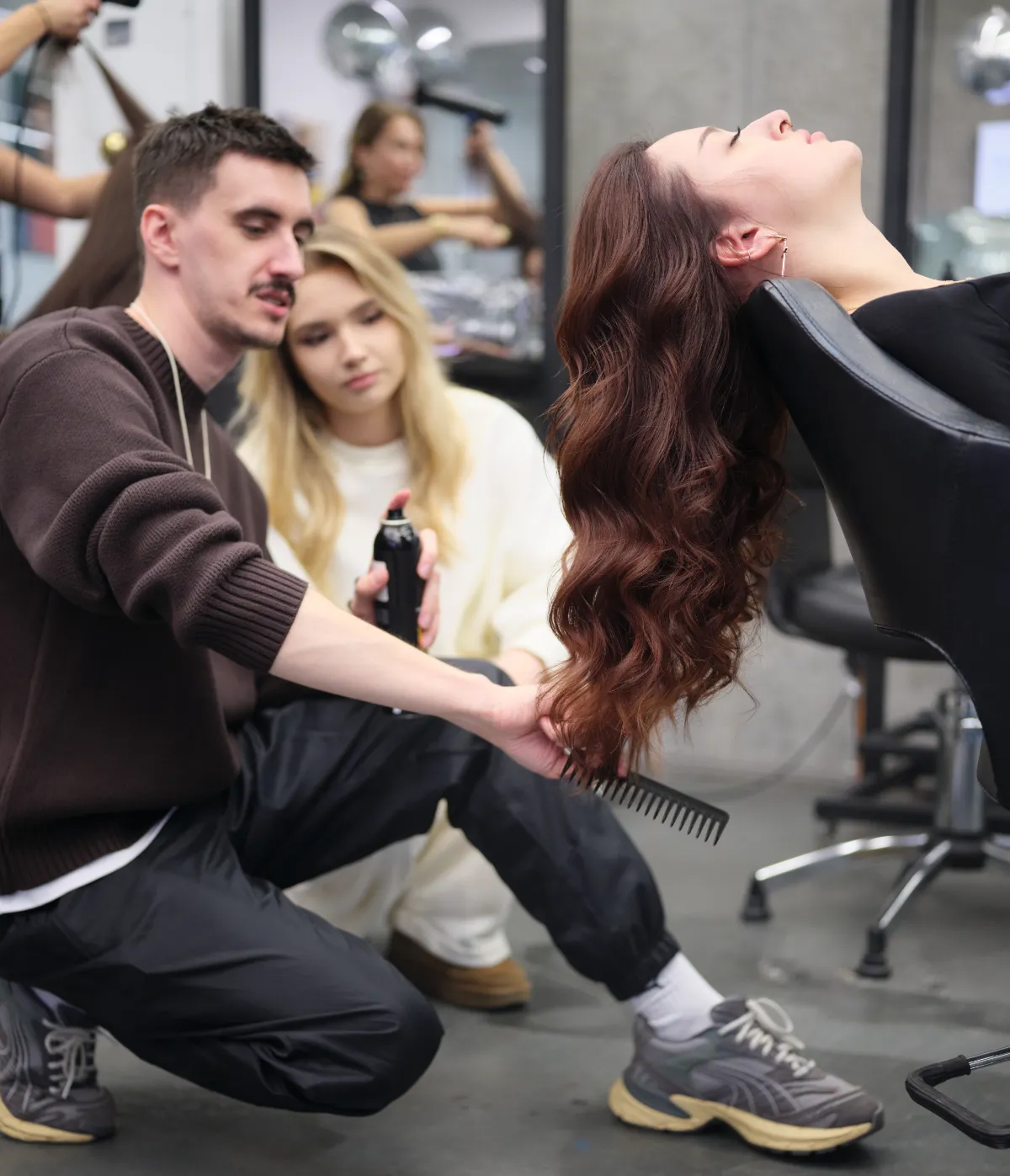 Майстер-перукар укладає довге руде волосся клієнтки: тримає пасма гребінцем і спреєм для фіксації, а поряд студент слідкує за його роботою для подальшого відпрацювання.