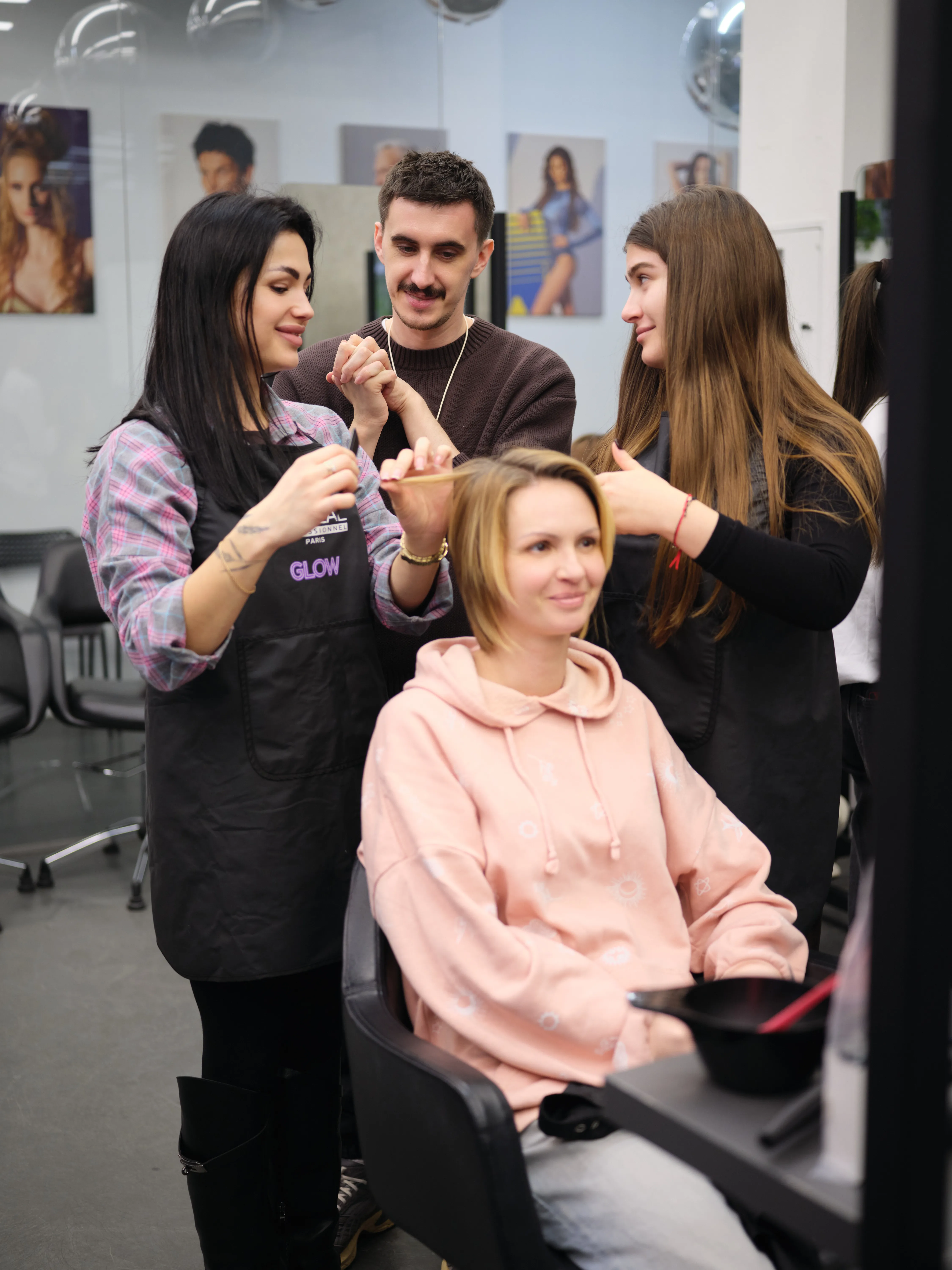 Студентки та викладач Blade Runner у салоні: молода жінка в рожевій худі сидить у кріслі, а майстрині в фартухах "GLOW" наносять фарбу на волосся за допомогою кисті та миски; поряд стоїть чоловік-викладач — демонстрація техніки фарбування блонду на занятті колористики.