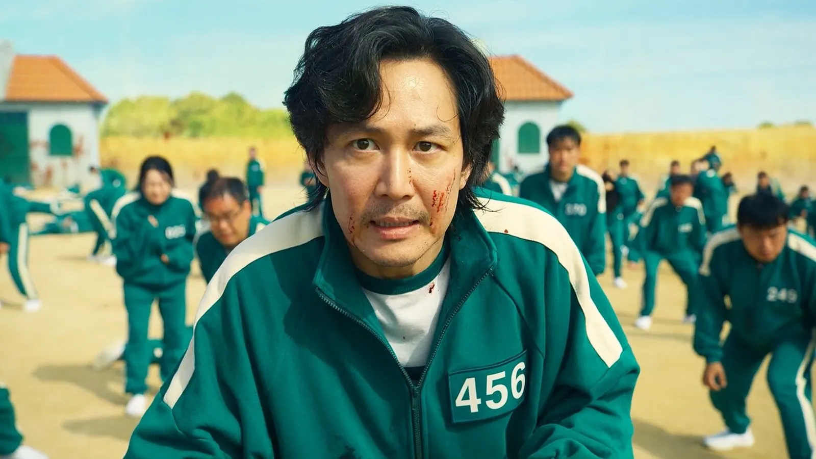 Сон Кі Хун (Гравець №456) у серіалі "Гра в кальмара", сезон 1: Лі Чон Че в зеленому костюмі з номером 456, з хвилястим чорним волоссям, закривавленим обличчям і напруженим виразом під час гри на тлі пустелі з іншими гравцями — драматичний момент для натхнення на зачіску з об'ємом.