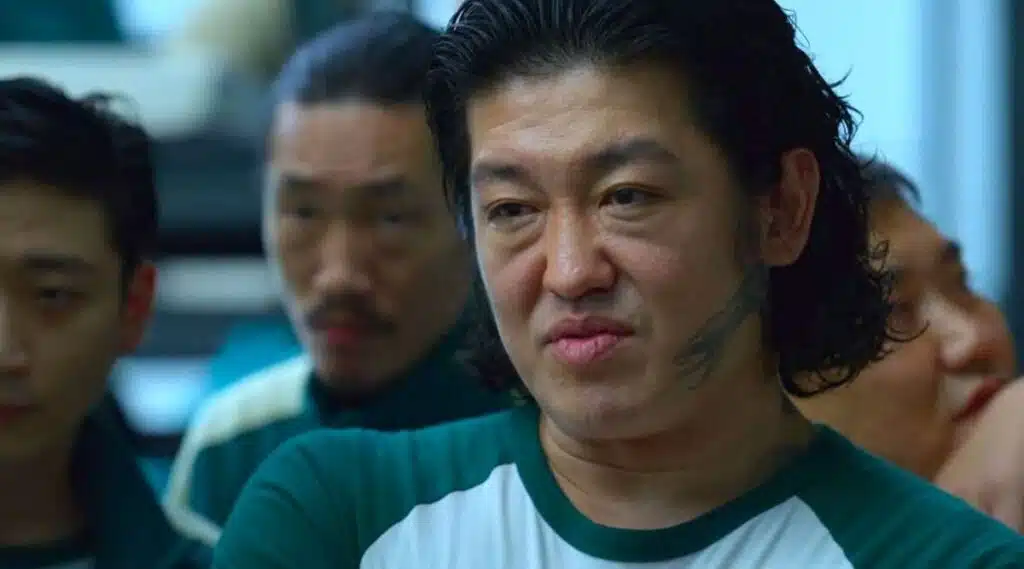 Чан Док Су (Гравець №101) у серіалі "Гра в кальмара", сезон 1, серія 2: чоловік з довгими хвилястими чорними локонами, зачесаними назад, у зеленому спортивному костюмі з номером 101, з татуюванням на обличчі та агресивним виразом — брутальний образ для косплею з акцентом на зачіску.