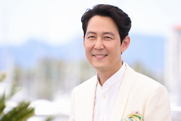 Лі Чон Че на Каннському фестивалі: актор у елегантному білому костюмі з білою сорочкою, з акуратною зачіскою "сайд парт" на чорному волоссі, посміхається на тлі моря та пальм — стильний повсякденний образ натхненний роллю Сон Кі Хуна.