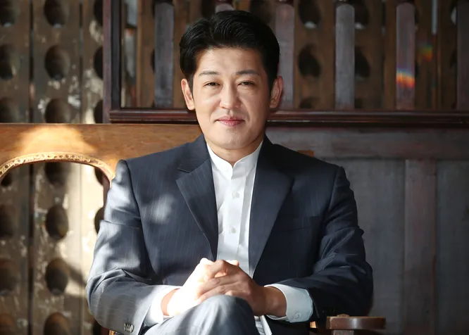 Хо Сон Тхе на фотосесії: актор у сірому костюмі з білою сорочкою сидить у дерев'яному кріслі з руками на колінах, з акуратною короткою зачіскою на чорному волоссі, посміхається на тлі дерев'яних панелей і бочок — елегантний образ для натхнення стилем