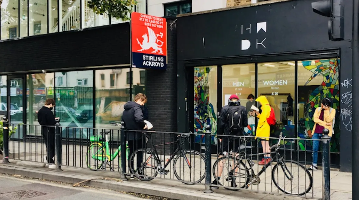 Зовнішній вигляд барбершопу Hurwundeki під залізничними арками в Лондоні: чорний фасад з логотипом HKDK, червоний прапор з драконом, люди в черзі біля велосипедів і стріт-арту — унікальний концепт барбер + корейський ресторан.