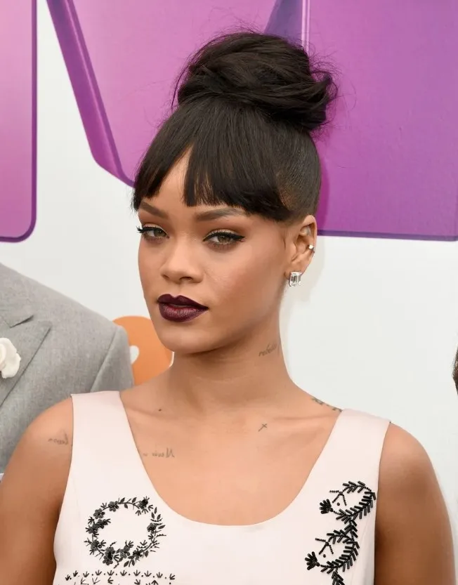 Ріанна на червоній доріжці з ефектним високим пучком у римському стилі на густе чорне волосся, прямими чубчиком, smokey-очима, бордовою помадою та татуюваннями на шиї; одягнена в рожевий топ з квітковою вишивкою, поруч чоловік у сірому костюмі, фіолетовий фон з логотипом MTV.