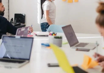 A meeting room table with computers on it and people around