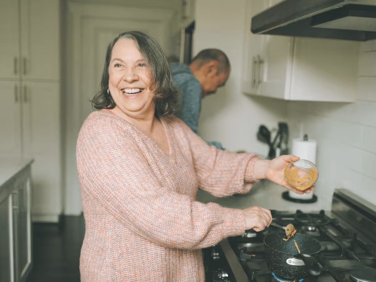 A joyful senior woman cooking in a modern kitchen, smiling warmly as she stirs a small pot on the stove.
