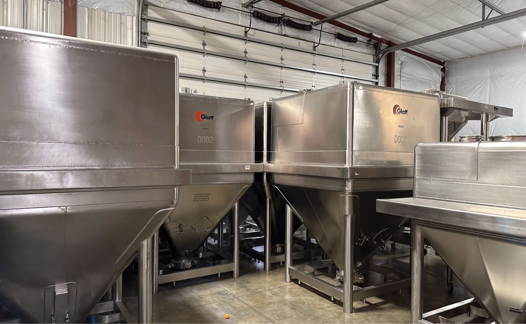 Industrial stainless steel Glatt processing equipment inside a warehouse with concrete floor and roll-up door.