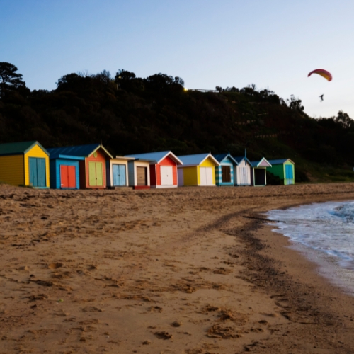 there are many beach huts on the beach with a parasail in the background