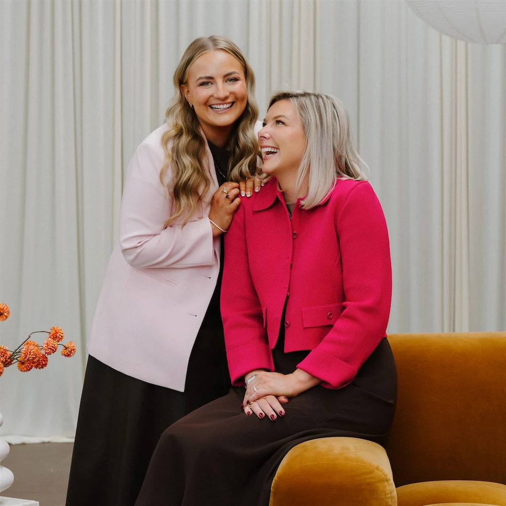 Two smiling women, one standing wearing a light pink blazer and the other seated wearing a bright pink jacket, posing in a room with beige curtains and an orange couch.