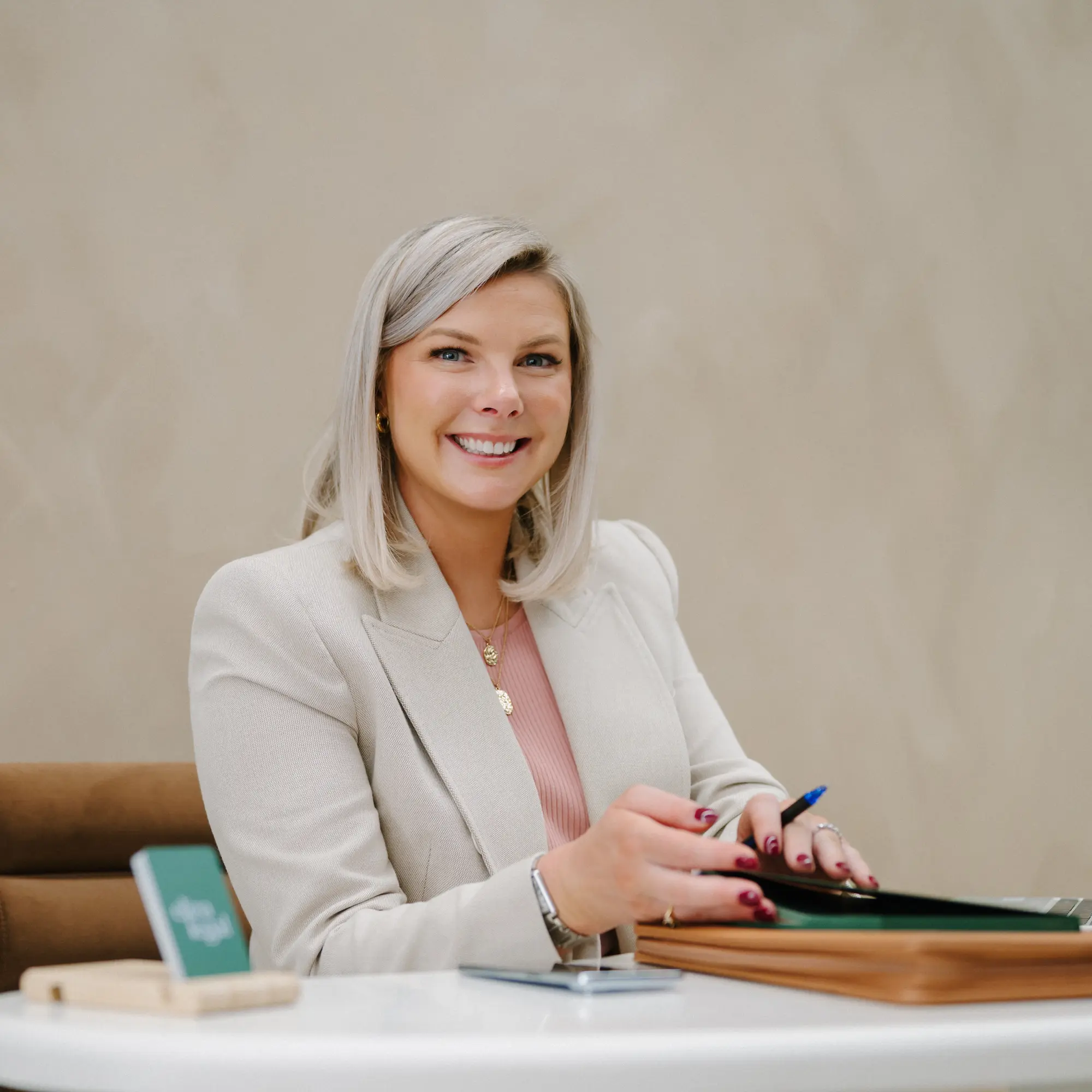 Smiling woman with blonde hair in a beige blazer sitting at a white table holding a pen and folder.