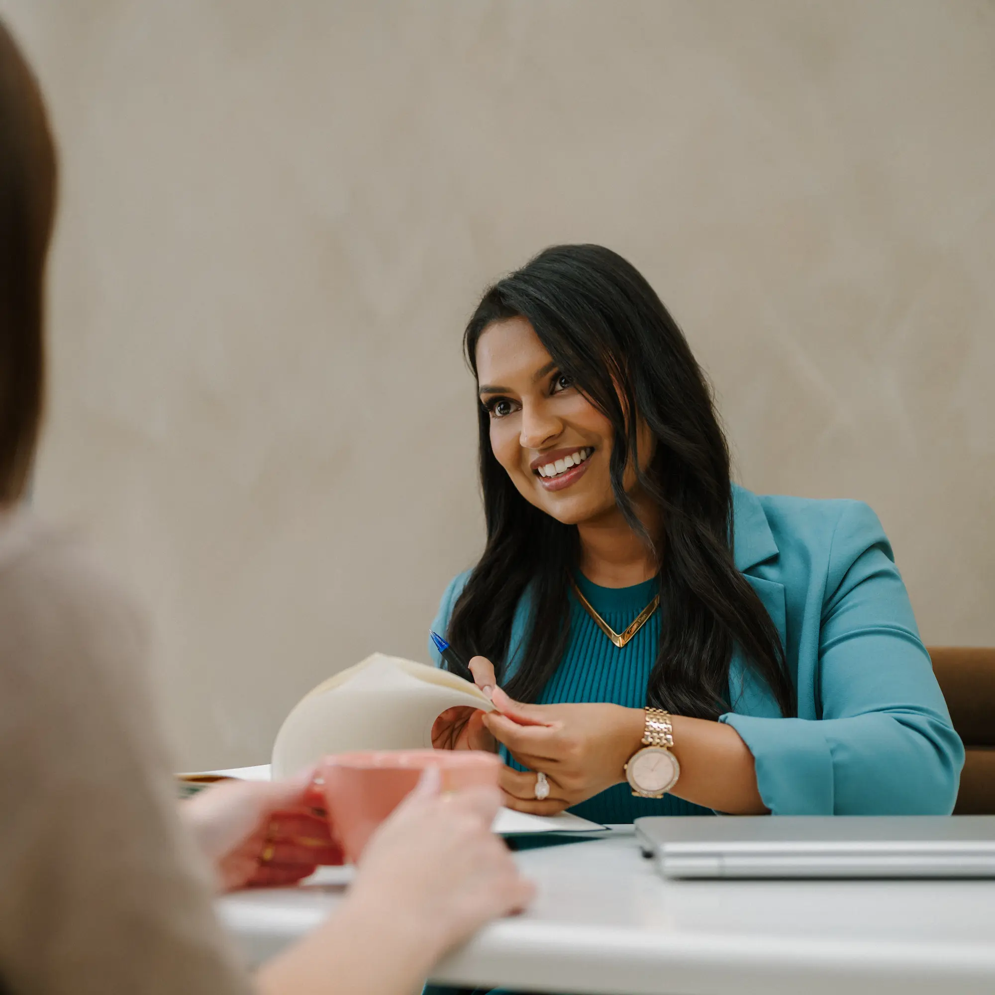 Smiling woman in teal blazer holding a pen and paper, talking to another person across the table.