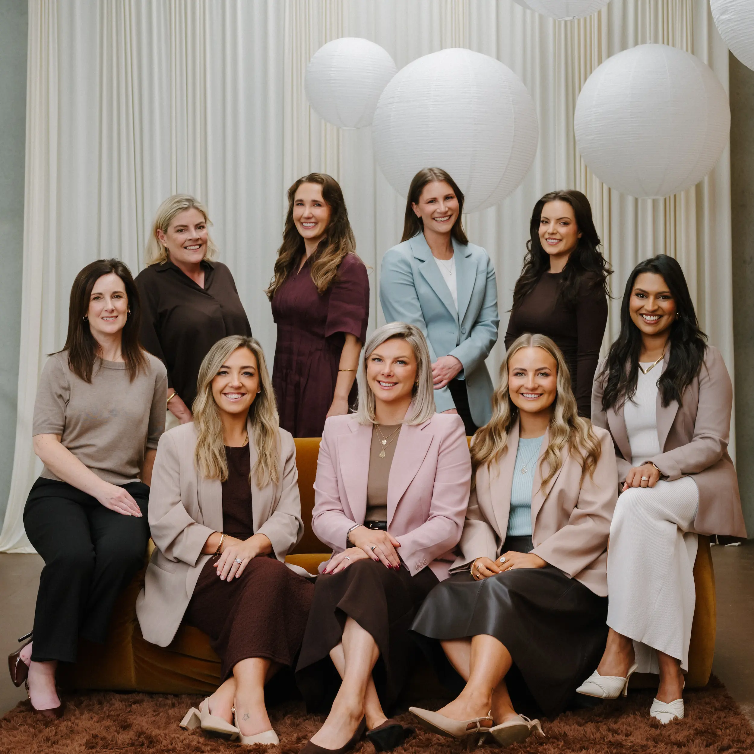 Group portrait of nine professional women sitting and standing indoors with white hanging paper lanterns in the background.