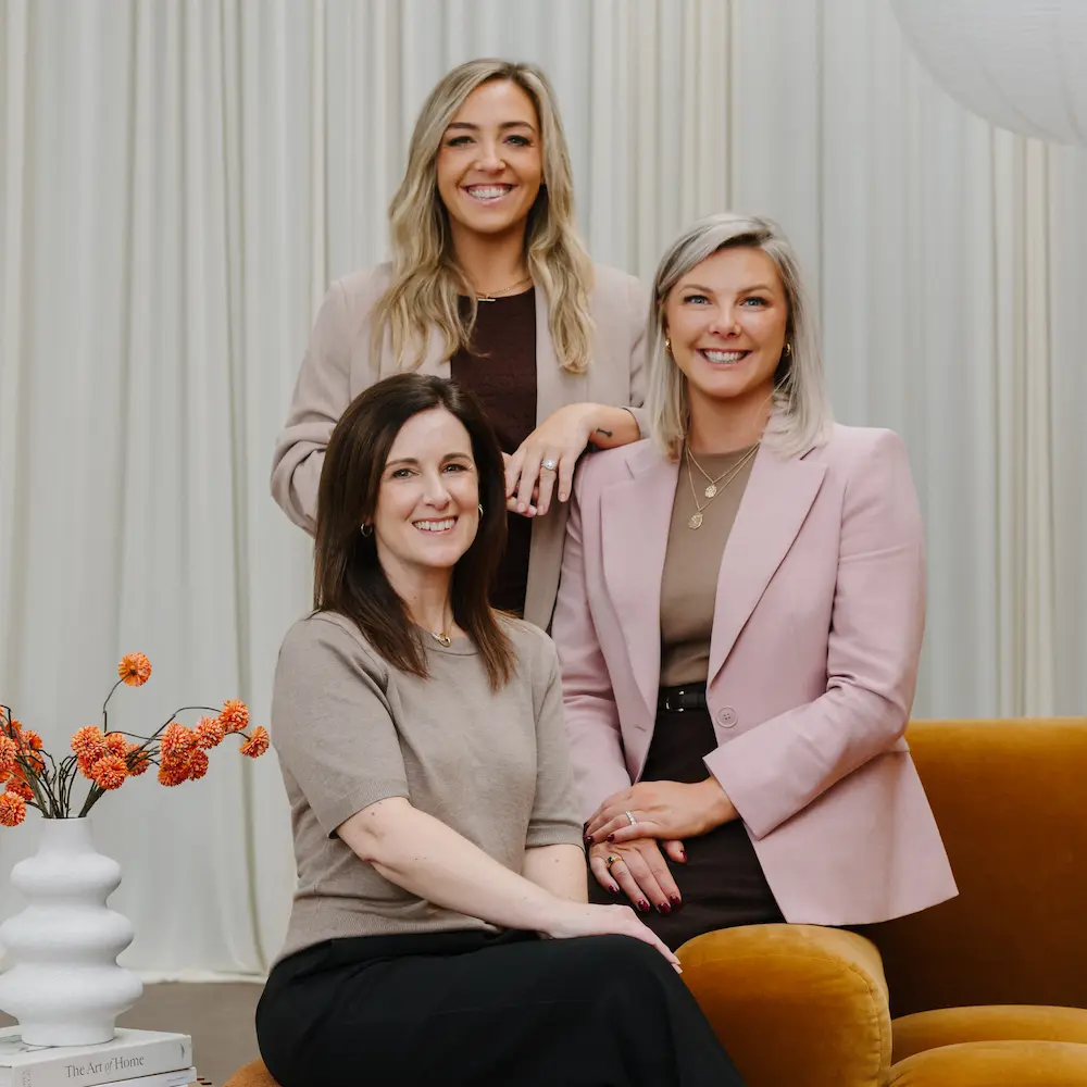 Three smiling women posing indoors with a white vase of orange flowers on a table.