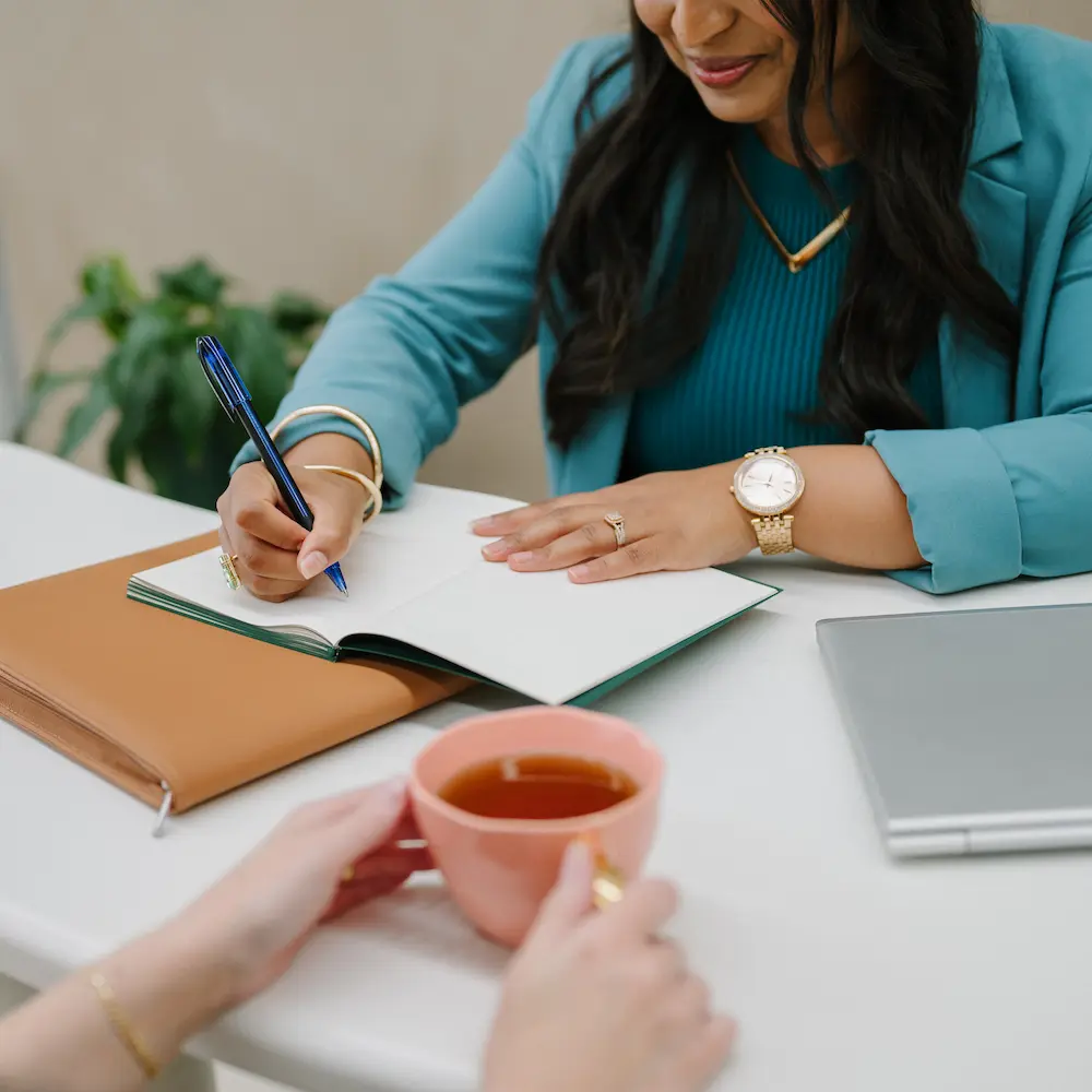 Woman in teal jacket writing in an open notebook at a white table with a tan folder and closed laptop nearby, and another person holding a pink cup of tea.