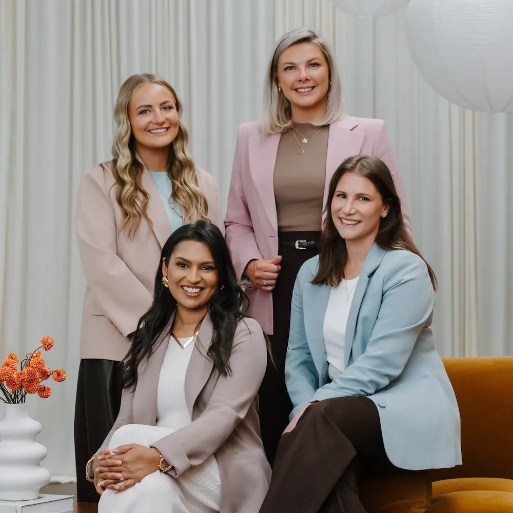 Portrait of four professional women smiling, three standing and one sitting on a brown chair, with a white vase and orange flowers on the side.