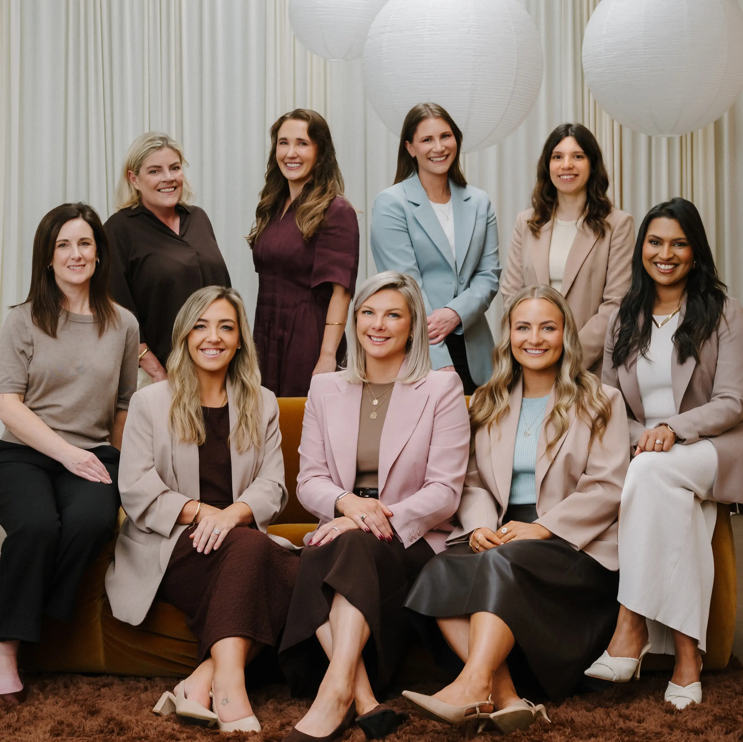 Seven women in smart casual attire smiling indoors on a brown couch and standing behind it.