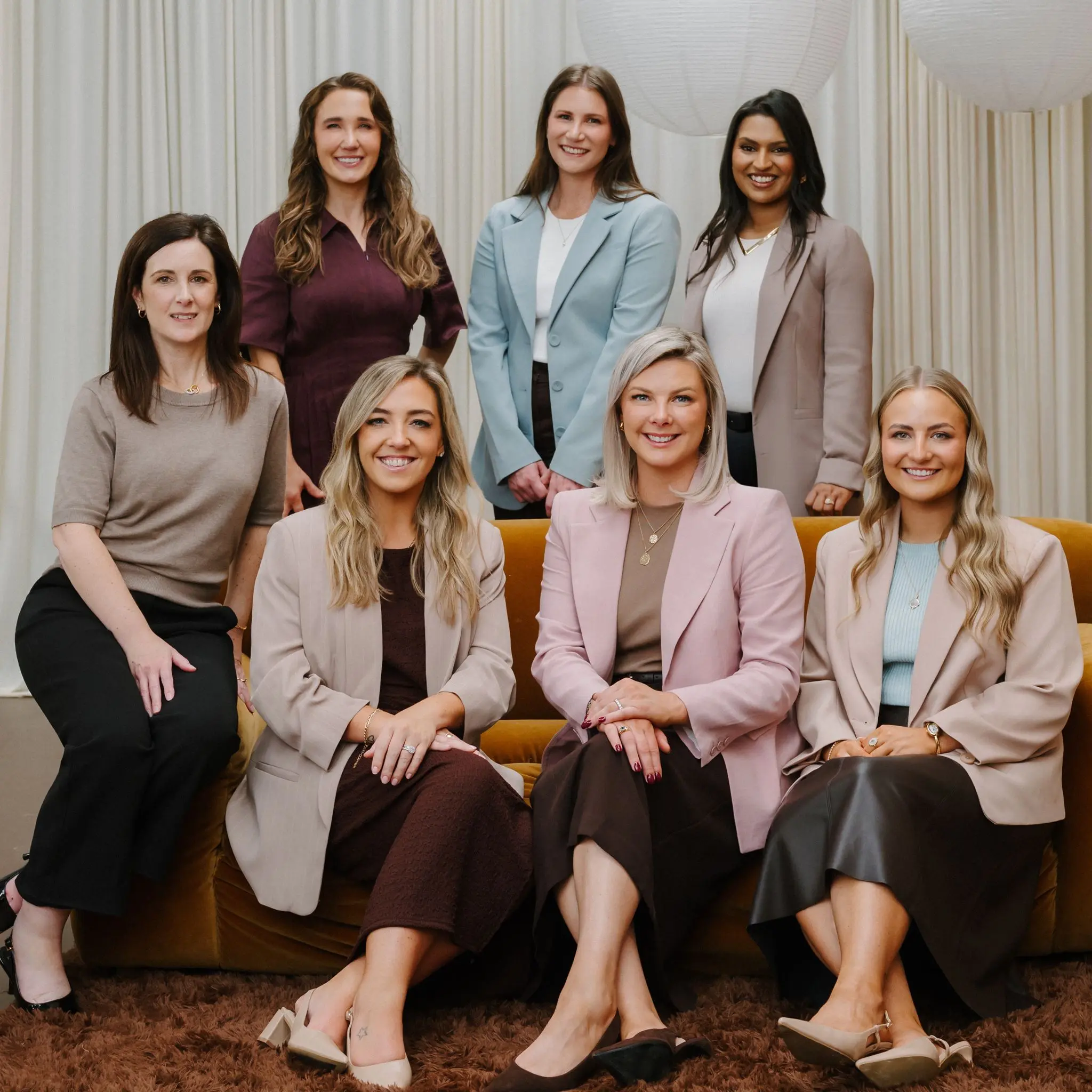 Seven women in smart casual attire smiling indoors on a brown couch and standing behind it.