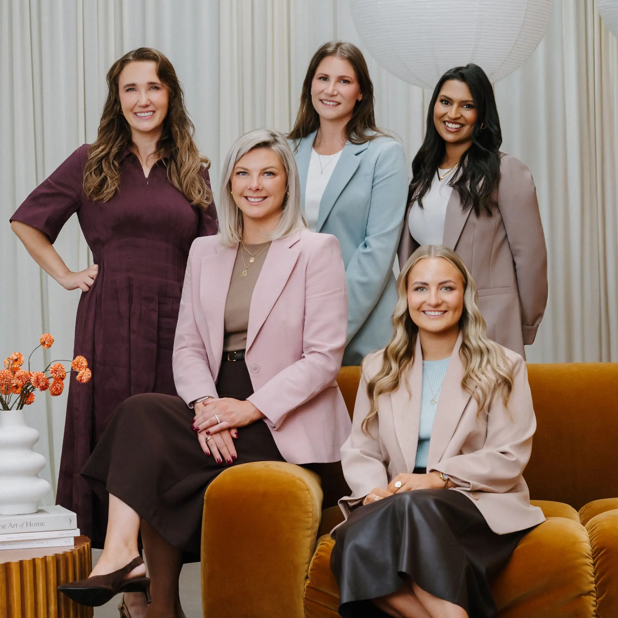 Portrait of five professional women smiling, three standing and two sitting on a brown chair, with a white vase and orange flowers on the side.