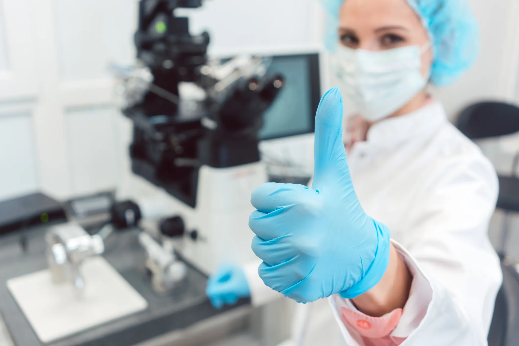Scientist in a mask and blue gloves giving a thumbs up beside a microscope in a laboratory