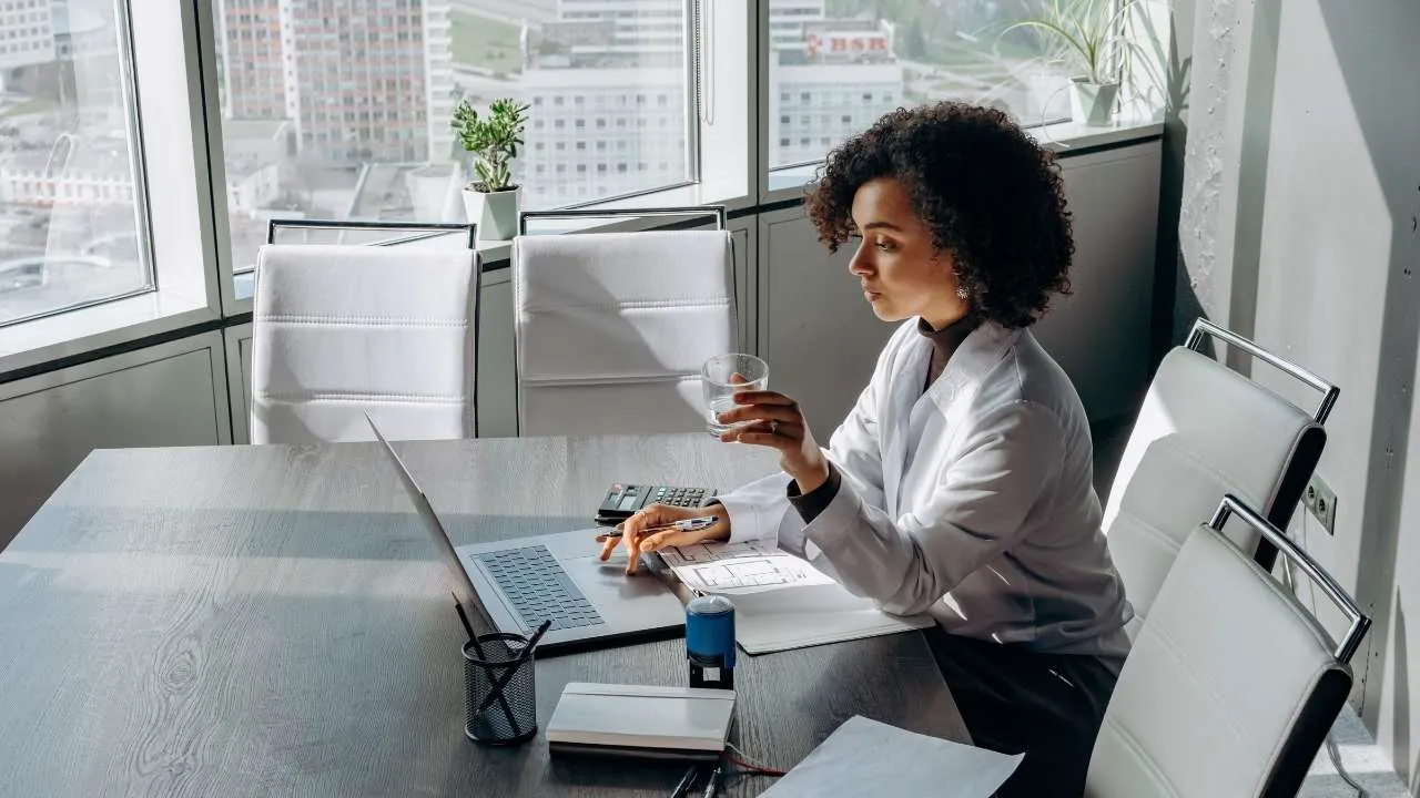 A professional dressed in white using a laptop at a desk and holding a glass of water