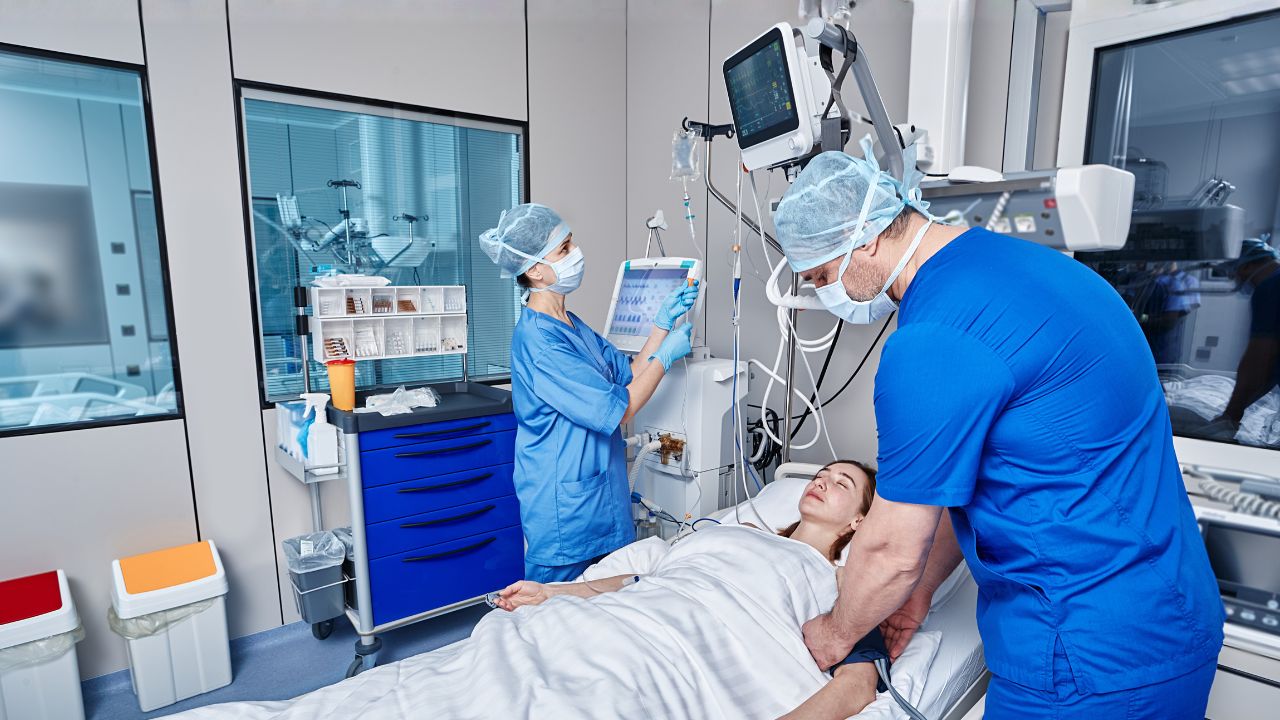 Two healthcare professionals monitor a patient lying in a hospital bed in a critical care unit