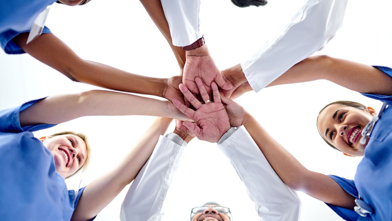 Healthcare team in scrubs and lab coats standing in a circle with hands stacked together