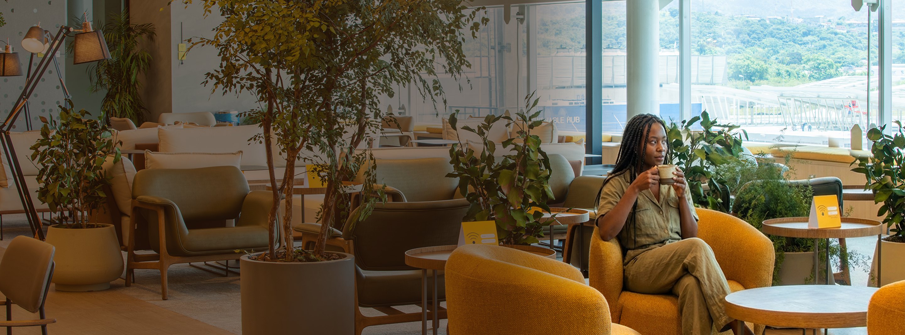 Área de convivência do lounge Nomad com poltronas amarelas, plantas e vista para o aeroporto