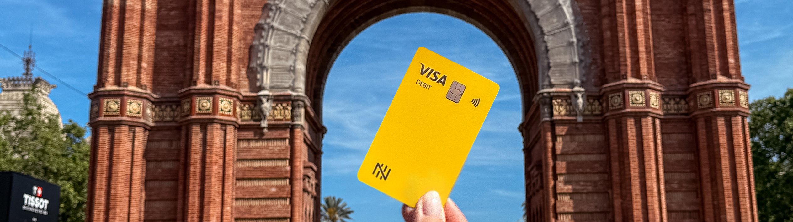 Mão segurando o cartão Nomad amarelo em frente ao Arco do Triunfo de Barcelona