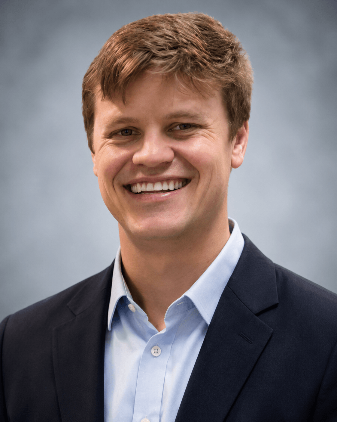 Smiling man with light brown hair wearing a light blue shirt and dark blazer on gray background.