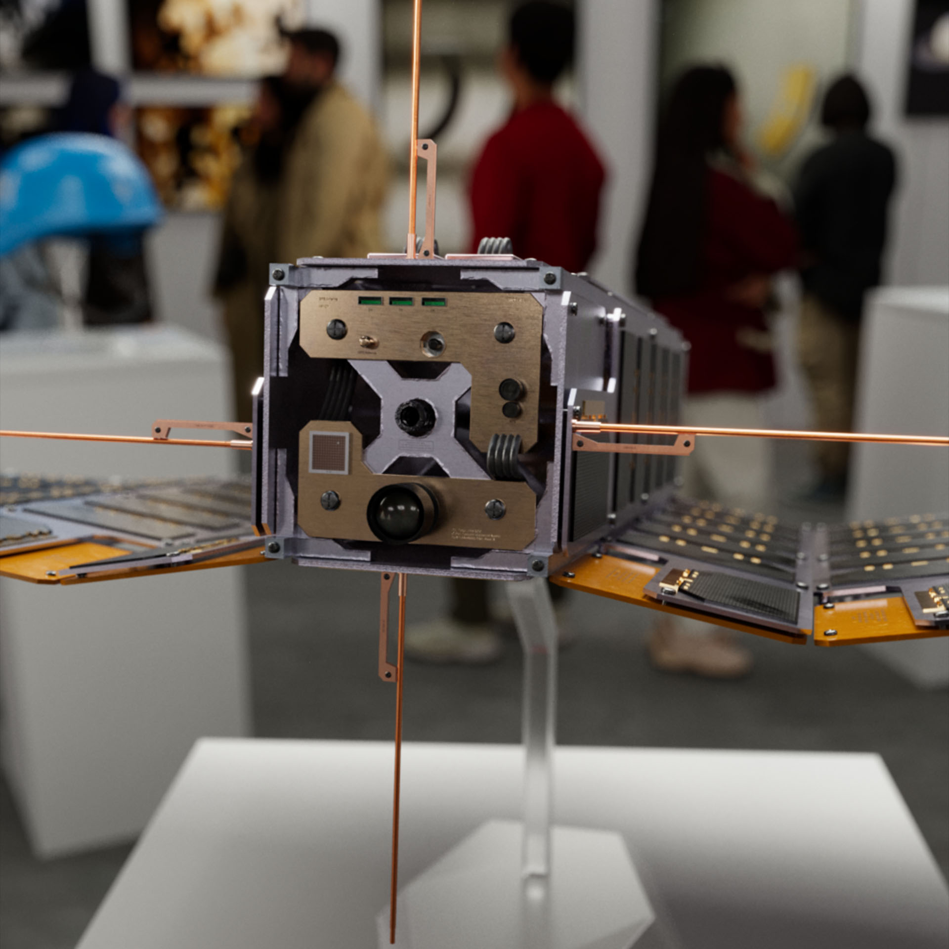 A military CubeSat displayed on a stand in an art gallery displaying other artifacts and art from Operation: Deli Platter.