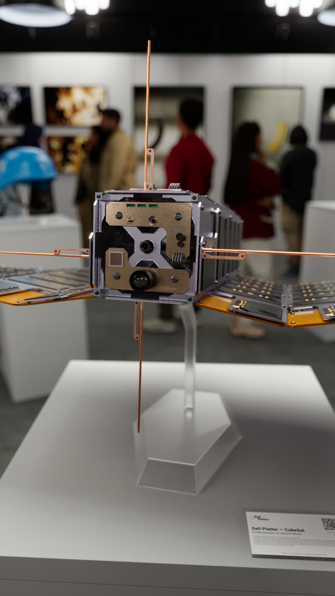 A military CubeSat displayed on a stand in an art gallery displaying other artifacts and art from Operation: Deli Platter.