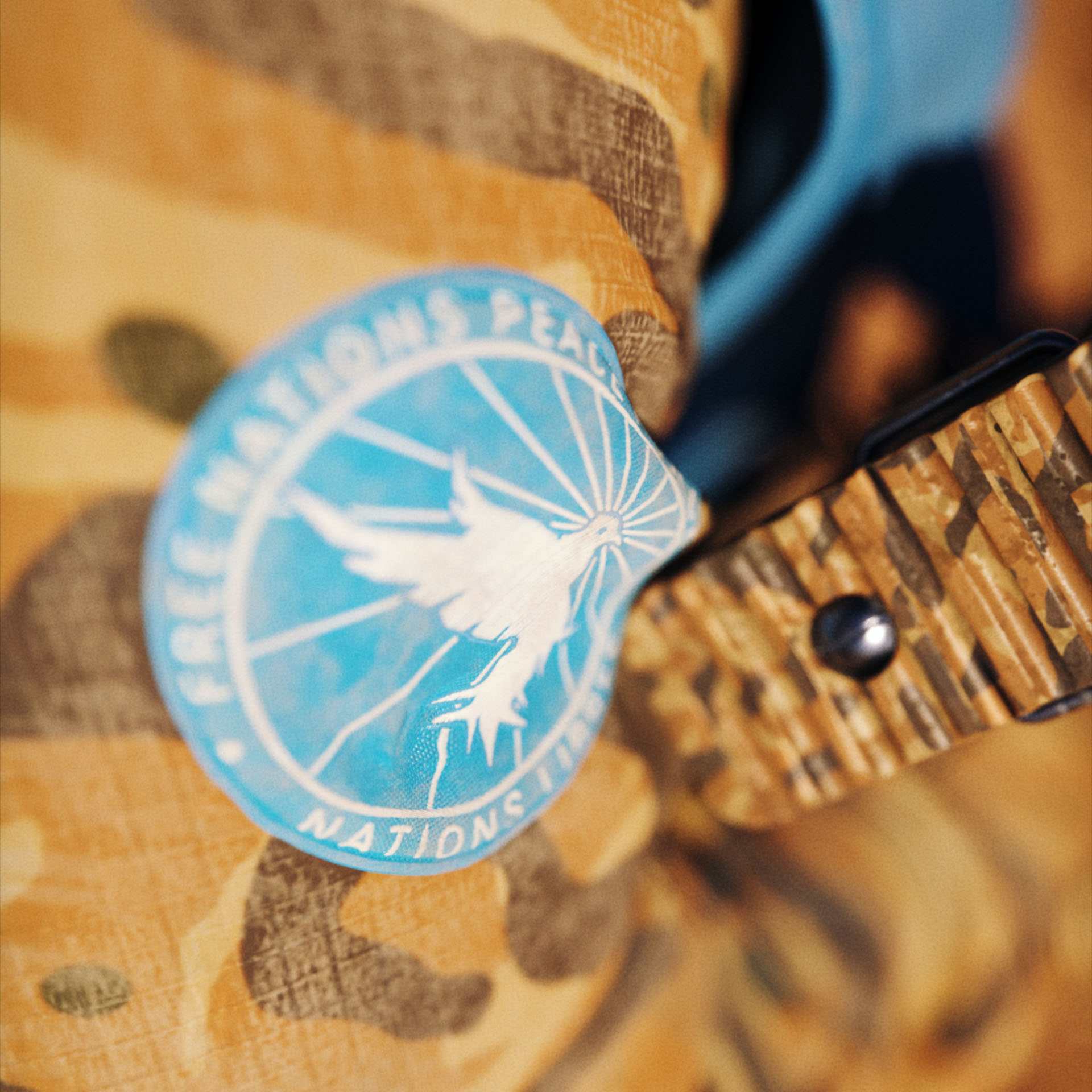 A close-up on a military fatigue with the Sand Root camouflage pattern and a bright blue military patch featuring a white dove with rays coming from it for the Free Nations Peace Force (FNPF) from Operation: Deli Platter.