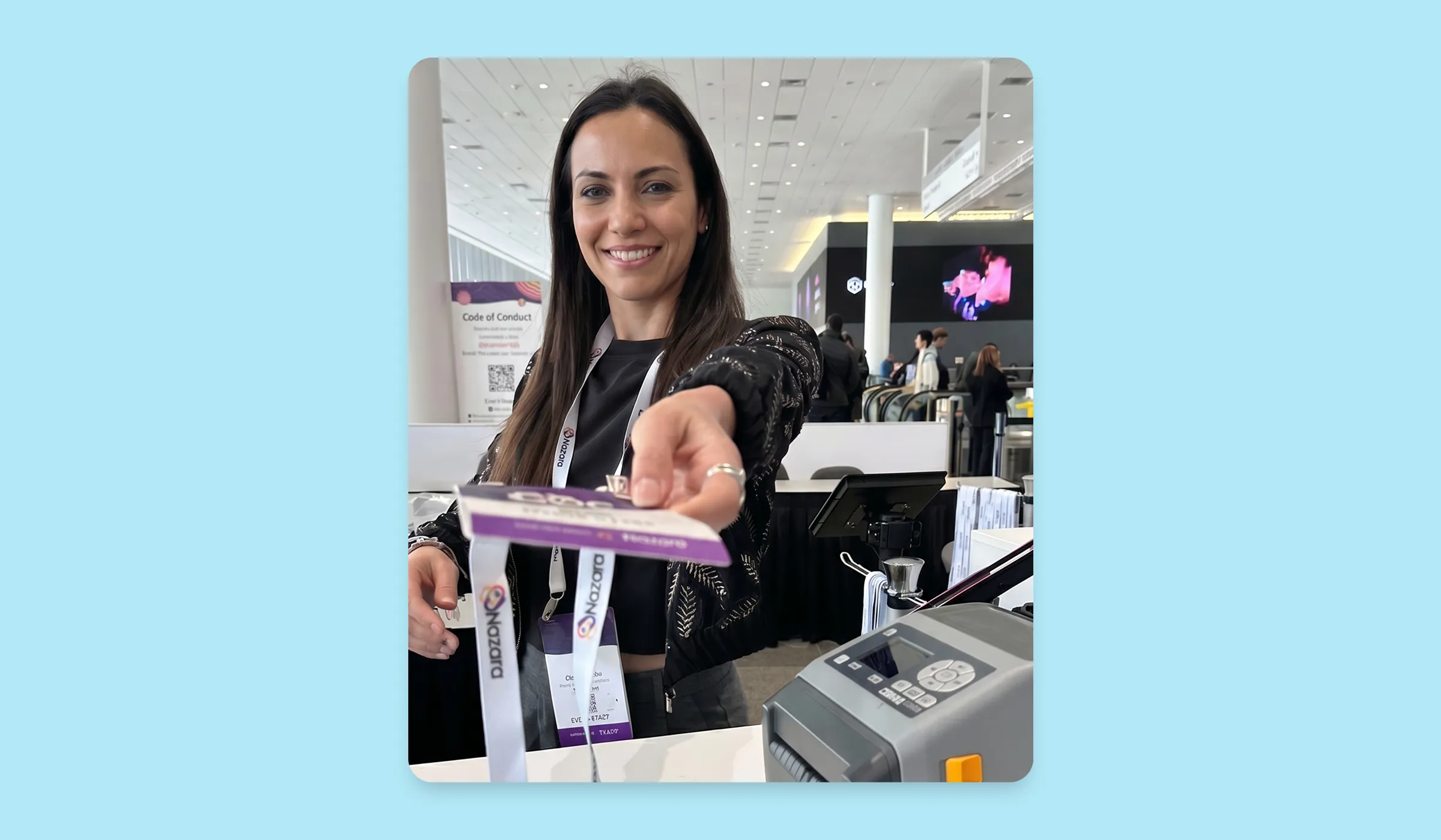 Smiling event staff member handing a badge or card to an attendee at a conference registration desk, with a busy exhibition hall in the background.