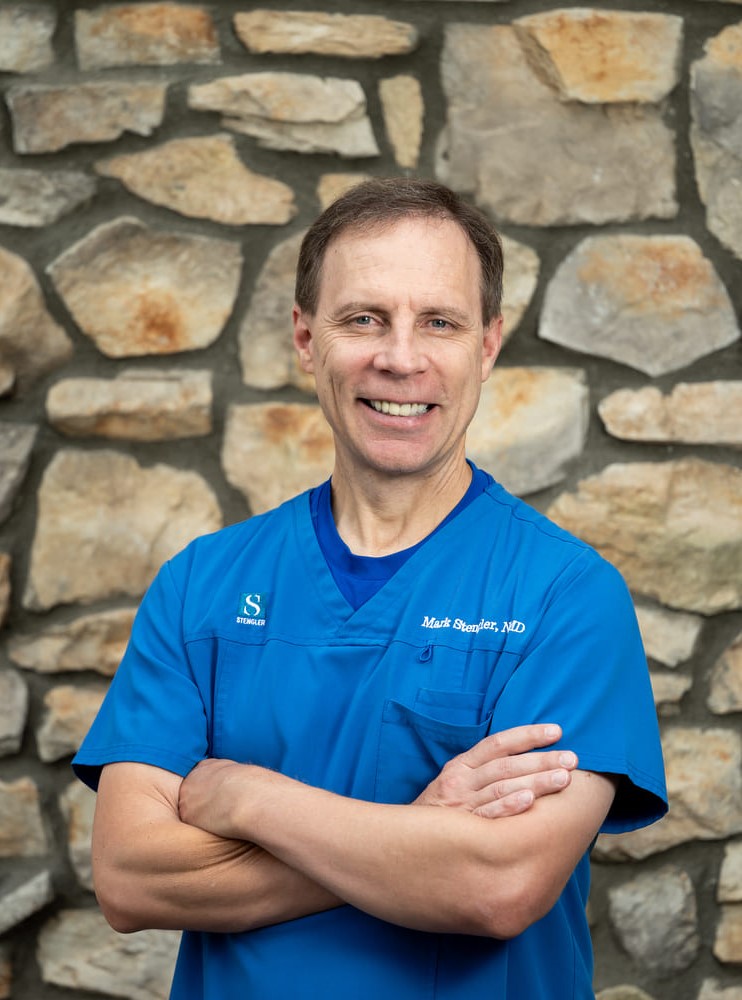 Mark Stengler Smiling wearing a white lab coat, blue shirt and stethoscope draped around his neck