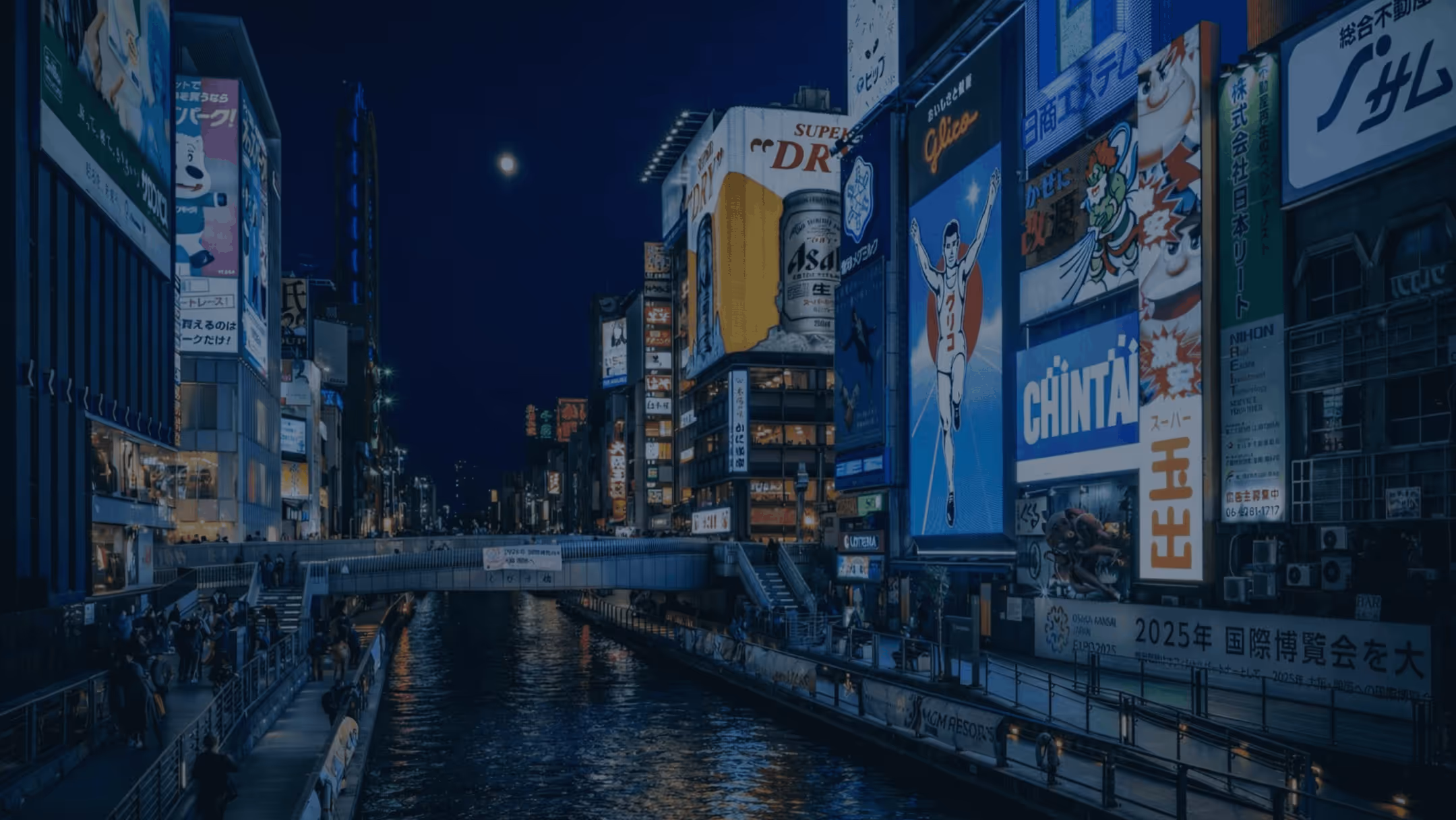 Nighttime cityscape of a canal lined with illuminated billboards and buildings, with people walking along the riverside paths.