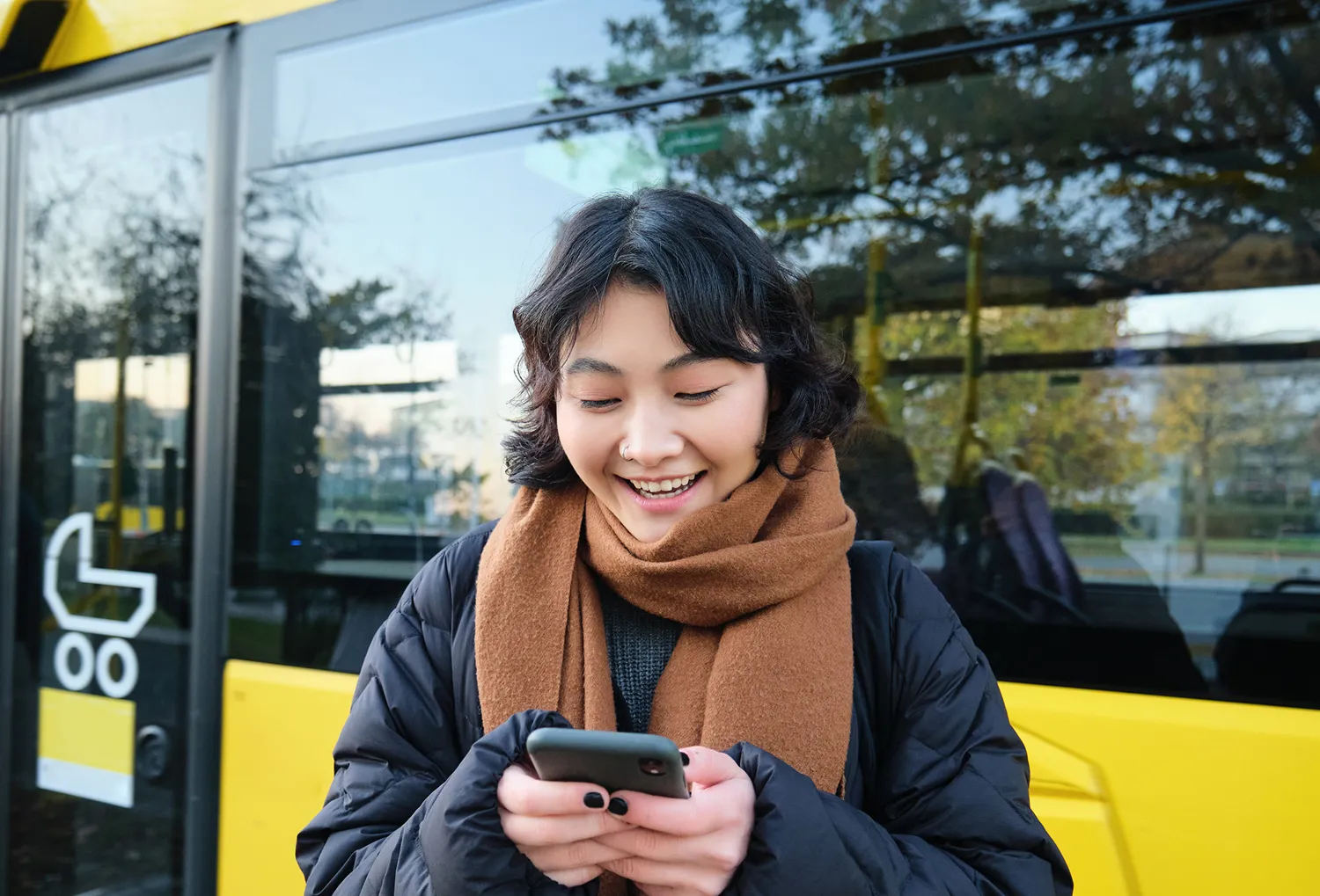 A woman standing in front of a bus looking at her cell phone.