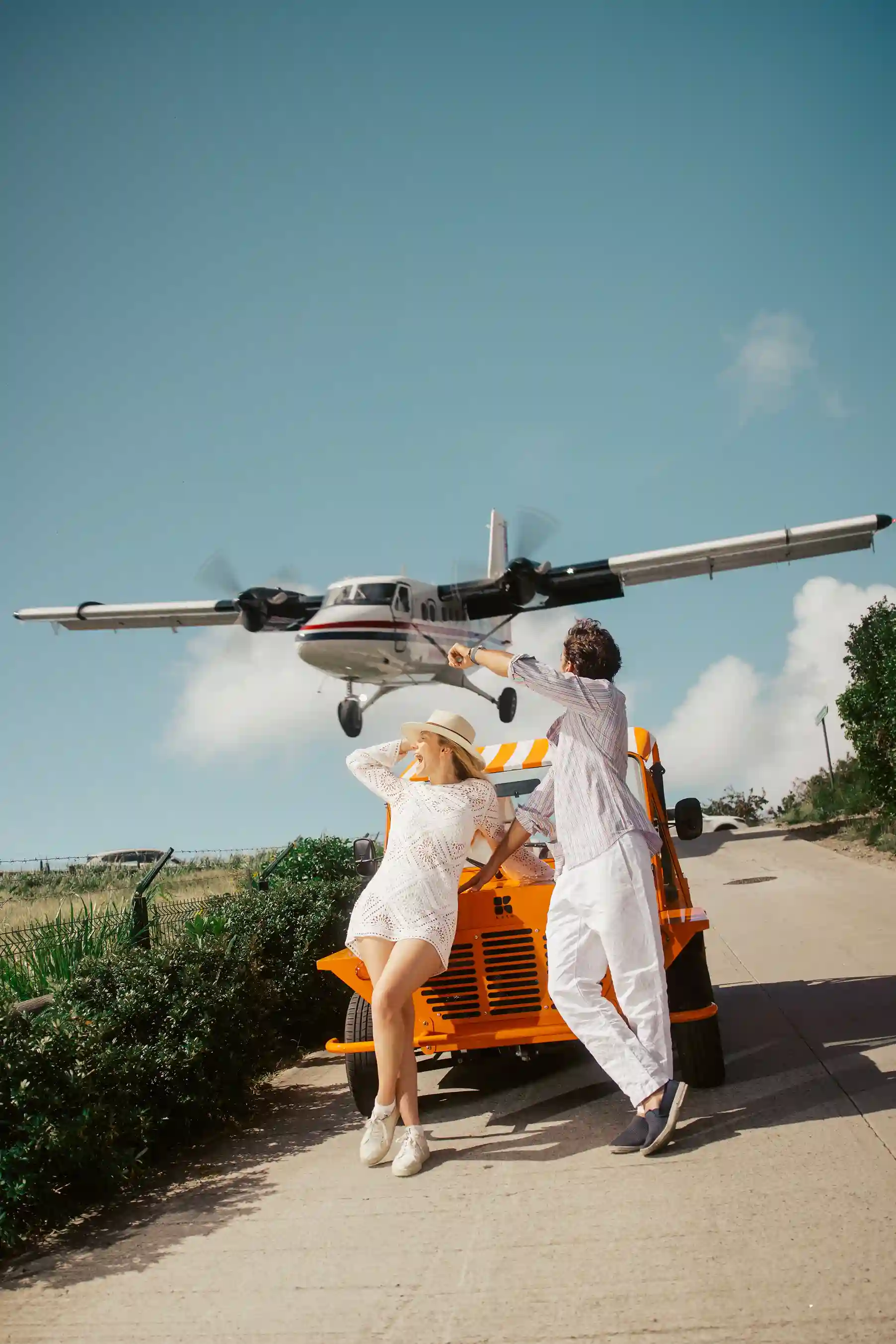 Man and woman leaning on an electric mini moke beach car from the brand Kate with a small plane flying low behind them.