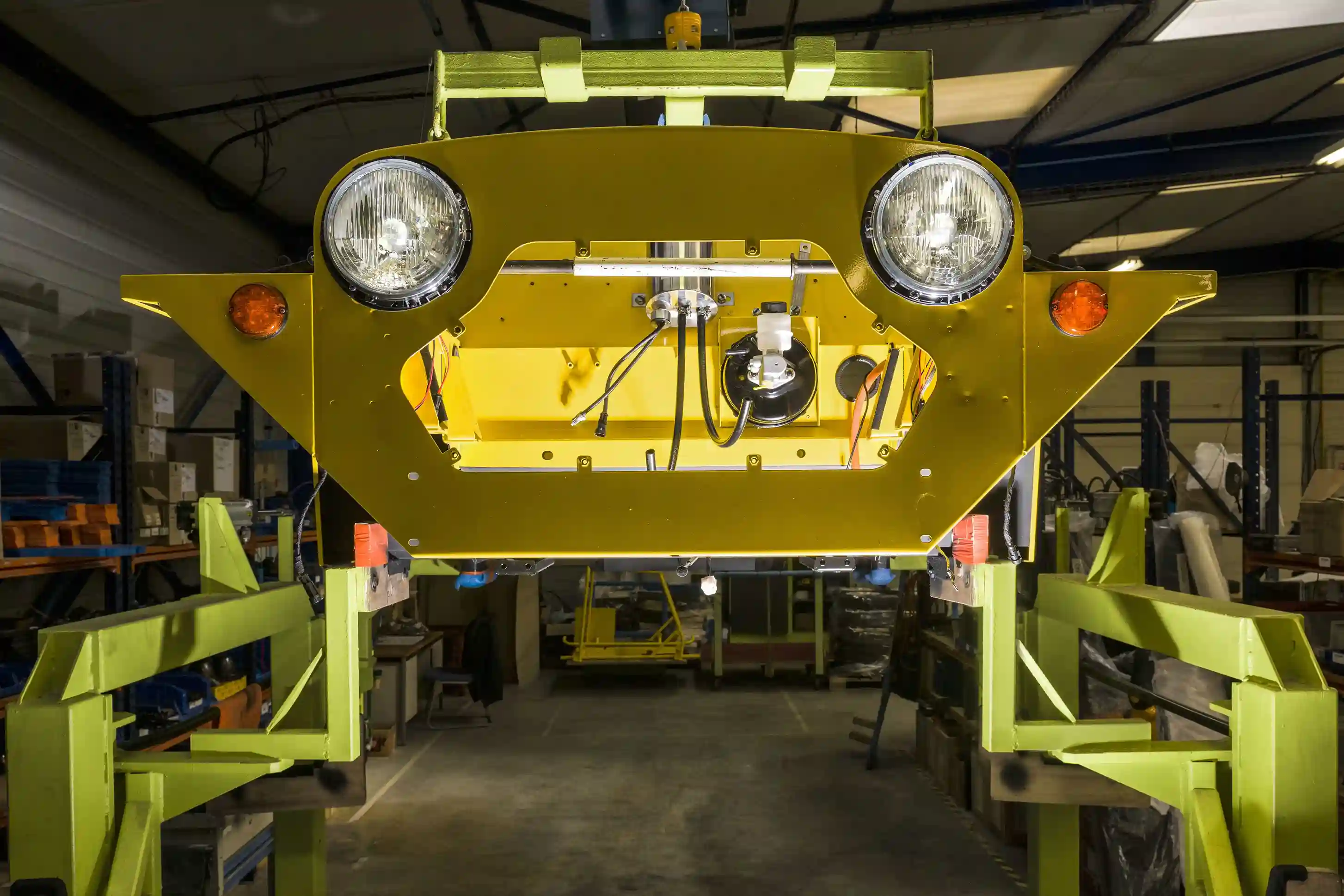 Front section of a yellow electric Mini Moke beach car Kate suspended on a green assembly line in a factory setting.