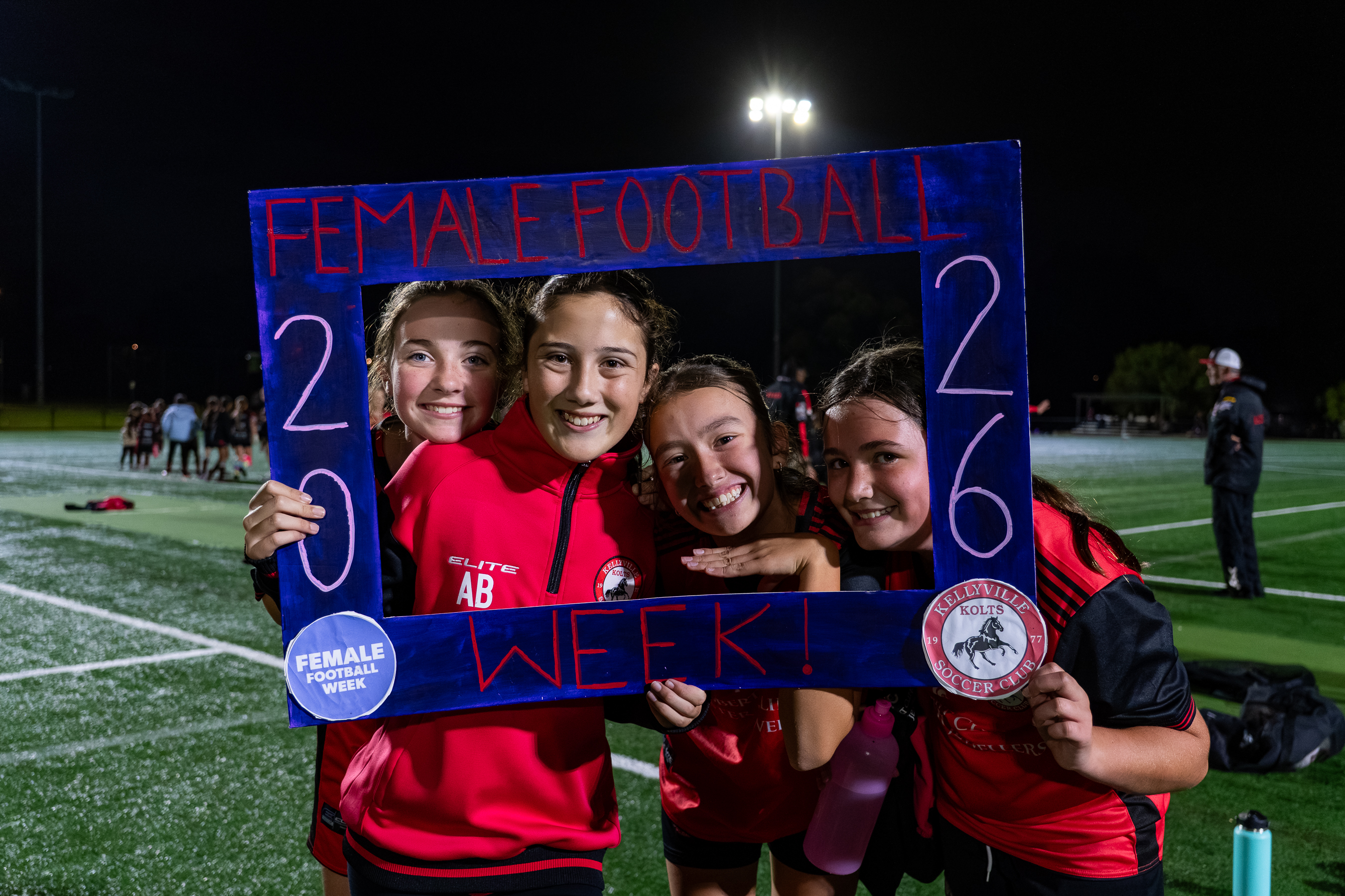Four smiling girls on a soccer field at night hold a blue frame that reads 'Female Football Week! 2026' with the Kellyville Kolts Soccer Club logos, celebrating female football.