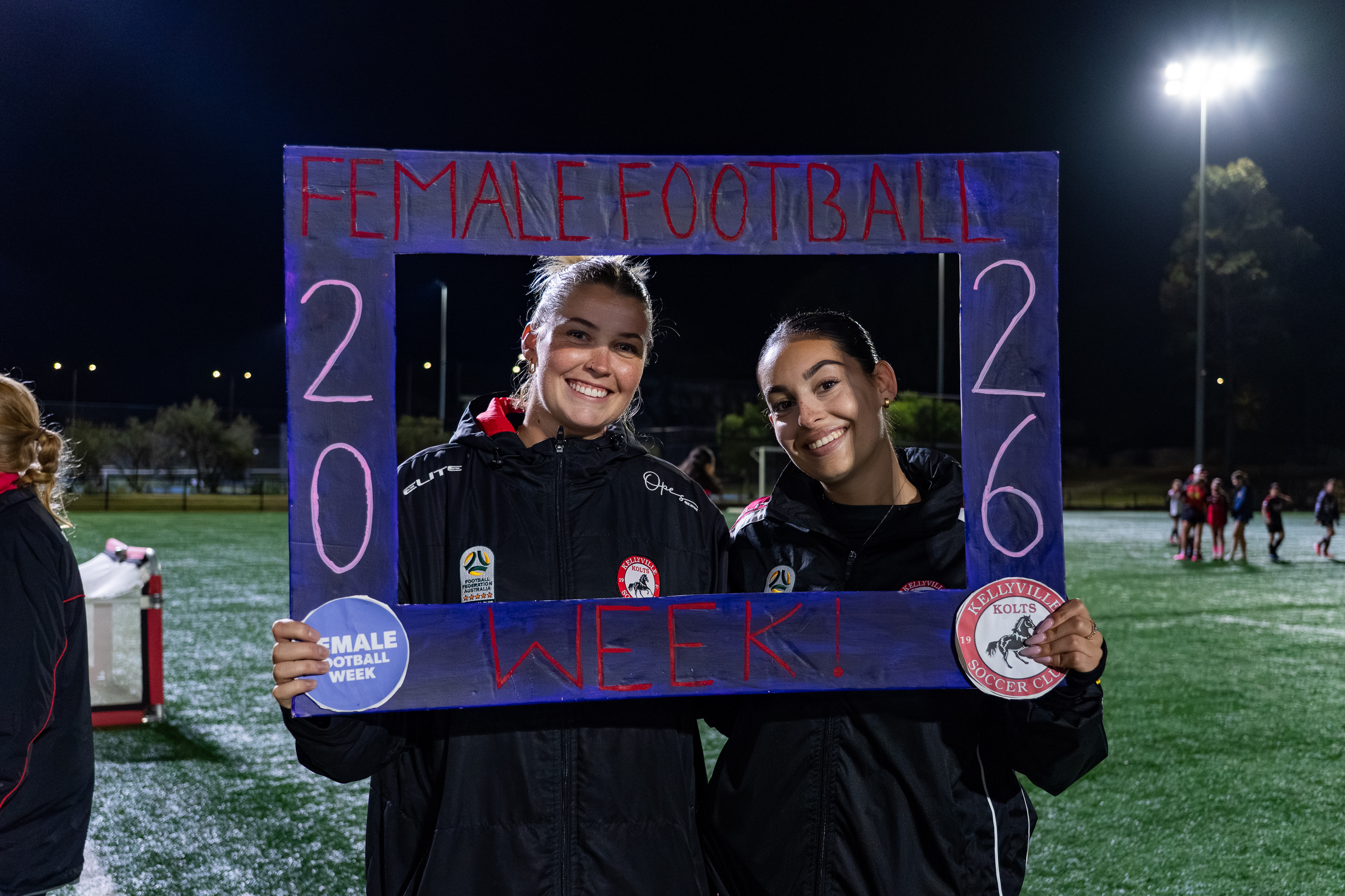 Two smiling women wearing Kellyville Kolts Soccer Club jackets hold a large blue frame that reads 'FEMALE FOOTBALL WEEK! 2026' while standing on an illuminated soccer field at night.