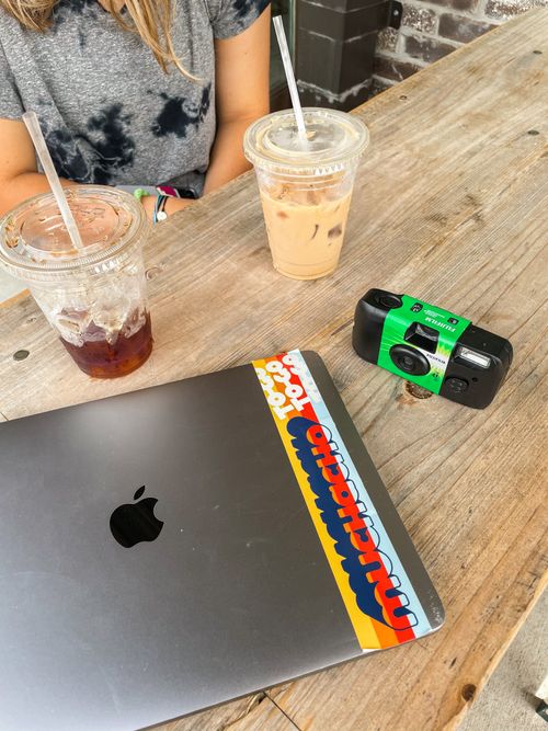 Disposable film camera, iced coffee, and computer on a wood table