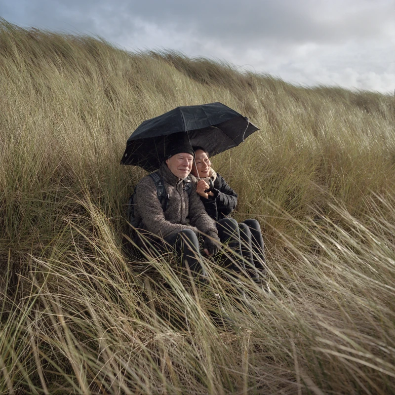 Une femme âgée portant des lunettes rouges est assise dans un fauteuil en bois près d'une fenêtre avec des rideaux sombres dans un salon encombré.