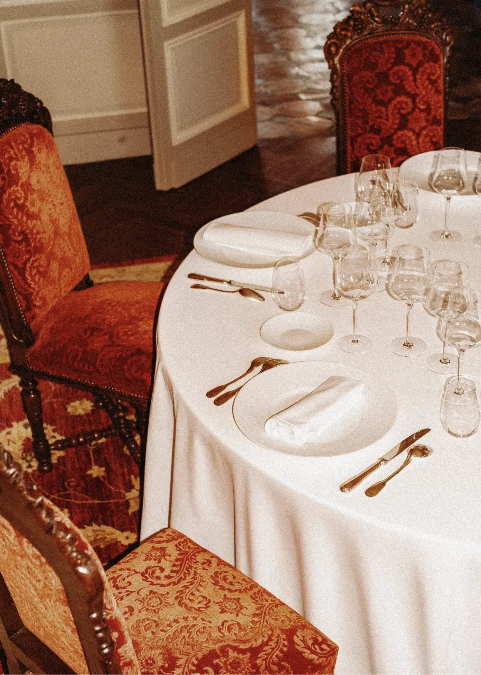 Table ronde dressée avec une nappe blanche, couverts délicats, plusieurs verres à vin, et chaises anciennes ornées de tissu rouge à motifs.
